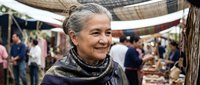 Senior Hispanic woman with gray hair smiling warmly at outdoor farmers market with colorful produce stalls and shoppers in background under fabric canopies.