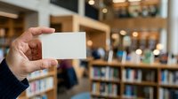 Person holding blank white business card in library setting with bookshelves and blurred background for branding and corporate identity mockups.