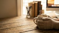 Cozy hands in knitted sweater holding warm coffee mug on rustic wooden table with books and stationery in soft morning light creating hygge atmosphere.