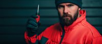Bearded man in red winter jacket and black beanie holding knife with threatening pose against dark background, dramatic cinematic lighting creates suspenseful atmosphere.