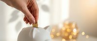 Hand inserting coin into white piggy bank with warm golden bokeh lights in background creating cozy savings and financial planning atmosphere.