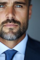 Close-up portrait of mature businessman with salt and pepper beard wearing blue suit and tie, expressing confidence and professionalism against blurred background.