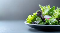 Fresh mixed green salad with lettuce leaves and sliced radish on dark plate against gray background, selective focus with copy space for menu design.