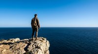 Male hiker with backpack standing on rocky cliff overlooking vast ocean and clear blue sky, adventure travel and exploration concept.