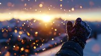 Winter gloved hand reaching towards golden sunset with falling snow and bokeh lights creating magical atmosphere against mountain landscape background.