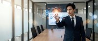 Asian businessman in dark suit touching holographic digital interface with network icons in modern conference room with glass walls and wooden table.