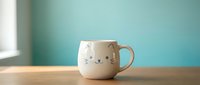 Cute white ceramic cat face mug with blue whiskers and sleepy eyes on wooden table against turquoise wall background for kitchen decor and morning coffee rituals.
