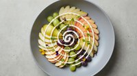 Colorful spiral arrangement of fresh sliced fruits on gray plate featuring apple, pear and other seasonal produce in artistic circular pattern for healthy eating.