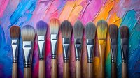 Row of clean professional art brushes arranged against vibrant multicolored acrylic paint texture background showing brush strokes in rainbow colors.