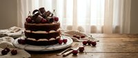 Decadent chocolate layer cake topped with fresh cherries and chocolate shavings on rustic wooden table. Homemade dessert for celebrations and special occasions.
