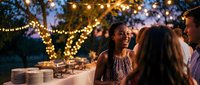 Young African American woman laughing joyfully at outdoor evening party with string lights and friends celebrating special occasion under trees.