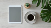 White tablet with blank screen on wooden desk with coffee cup, small succulent plant, and monstera leaves for modern workspace styling.
