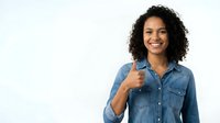 Young woman with curly dark hair and medium skin tone wearing denim shirt, giving thumbs up gesture, smiling at camera against white background for approval and positive feedback concepts.