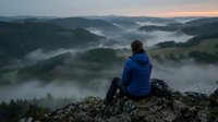 Young adult male, Caucasian, wearing blue winter jacket, sitting on rocky mountain peak overlooking misty forested valley at dawn, contemplating scenic landscape with fog-covered hills and evergreen trees.