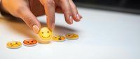 Hand selecting happy yellow emoji from row of emotion faces on white surface for customer feedback and satisfaction rating concept.