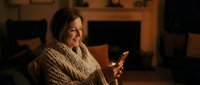 Middle-aged woman with brown hair wearing a cozy knit sweater, holding a smartphone while sitting indoors in warm lighting. Suitable for technology, communication, and lifestyle content.