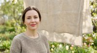 Young Caucasian woman in beige knit sweater smiling outdoors near stone wall with green garden background for lifestyle and wellness content.