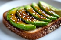Fresh sliced avocado on dark toasted bread garnished with honey drizzle and white sesame seeds, served on white plate. Close up macro food photography.