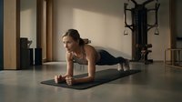 Athletic woman performing plank exercise on yoga mat in modern home gym with natural lighting and fitness equipment in background.