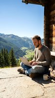 Young Caucasian man working on laptop outdoors at mountain cabin terrace with alpine valley view, remote work lifestyle and digital nomad concept.