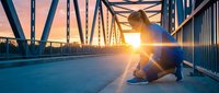 A young adult male in athletic wear crouches on a modern bridge at sunrise, silhouetted against golden sunlight streaming through steel framework, capturing motivation and urban fitness.