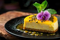 Passion fruit cheesecake slice garnished with fresh mint leaves and purple edible flower on black plate, passion fruit sauce and black sesame seeds. Food photography.