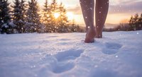 Barefoot woman walking through fresh snow at golden sunset with falling snowflakes creating magical winter atmosphere and peaceful outdoor scene.