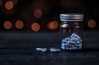 Glass jar filled with blue glowing capsules on dark surface against blurred bokeh lights background, creating mysterious medical or pharmaceutical atmosphere.