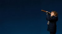 Young boy with curly hair wearing dark jacket and striped shirt, looking through telescope against deep blue background. Suitable for education, astronomy, science, and children's learning concepts.