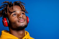 Young African American man with dreadlocks wearing yellow hoodie and red headphones looking up with hopeful expression against vibrant blue background.