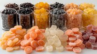 Colorful assortment of gummy candies and jelly beans in glass jars and scattered on white surface, featuring orange, yellow, black, and pink sweet treats in organized display.