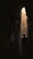 Silhouette of male explorer standing in dramatic cave with golden sunlight beam streaming through opening above creating atmospheric underground scene.