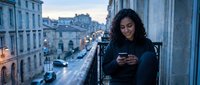 Young woman with dark curly hair and olive complexion, wearing dark clothing, checking smartphone on urban balcony overlooking European city street at dusk. Suitable for lifestyle, technology, and travel content.