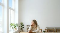 Woman in beige shirt sitting at desk by window with coffee cup, plants, and office supplies, suitable for business, work-from-home, and professional lifestyle content.