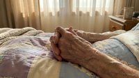 Elderly hands clasped together in prayer or meditation on hospital bed with soft morning light streaming through window curtains creating peaceful atmosphere.