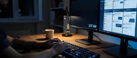Professional workspace with dual monitors displaying data analytics dashboard in dimly lit office environment with coffee cup and keyboard setup.