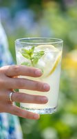 Person holding refreshing lemonade with fresh mint and lemon slices in glass outdoors on sunny day with blurred green garden background.