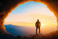 Silhouette of male hiker standing in mountain cave entrance at sunset, dramatic orange sky and mountain ranges creating inspiring outdoor scene.
