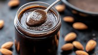 Homemade dark chocolate almond butter in glass jar with spoon and scattered nuts on black surface, creamy smooth texture with rich glossy appearance.