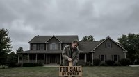 Sad homeowner man sitting behind for sale by owner sign in front of large suburban house on overcast day during housing market crisis.