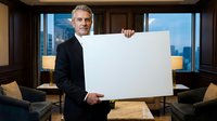 Confident mature Caucasian businessman in dark suit holding blank white sign in luxury office with city skyline view for corporate presentations and marketing.