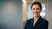 Mature woman with dark hair and fair complexion wearing navy blazer smiling at camera in modern office corridor with blurred background, suitable for corporate, business, and professional portraits.