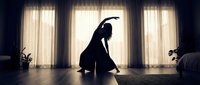 Woman practicing yoga in silhouette against bright window light, stretching in peaceful morning routine at home with natural sunlight streaming through curtains.