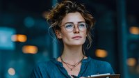Young professional woman with glasses and messy bun hairstyle in urban evening setting with bokeh lights background for business and lifestyle concepts.