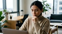 Focused young woman with dark hair and medium skin tone working on laptop in modern office space with plants and natural light.