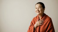 Mature woman with East Asian ethnicity wearing rust-orange shirt, hand on chest, smiling warmly against neutral background. Suitable for healthcare, wellness, gratitude, and lifestyle editorial use.