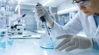 Female scientist in white lab coat and safety glasses conducting research experiment with pipette and petri dish in modern laboratory setting.
