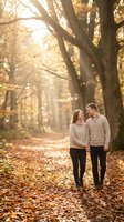 Young couple holding hands walking through golden autumn tree-lined path covered in fallen leaves. Warm sunlight filters through canopy creating romantic atmosphere for engagement, relationship, or lifestyle content.