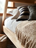 Cozy bedroom interior with wooden bed frame, white bedding, knitted beige throw blanket and navy striped decorative pillows in natural morning light.