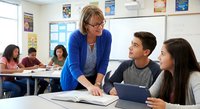 Middle-aged Caucasian female teacher in blue cardigan helping teenage students with schoolwork in modern classroom setting with educational posters.